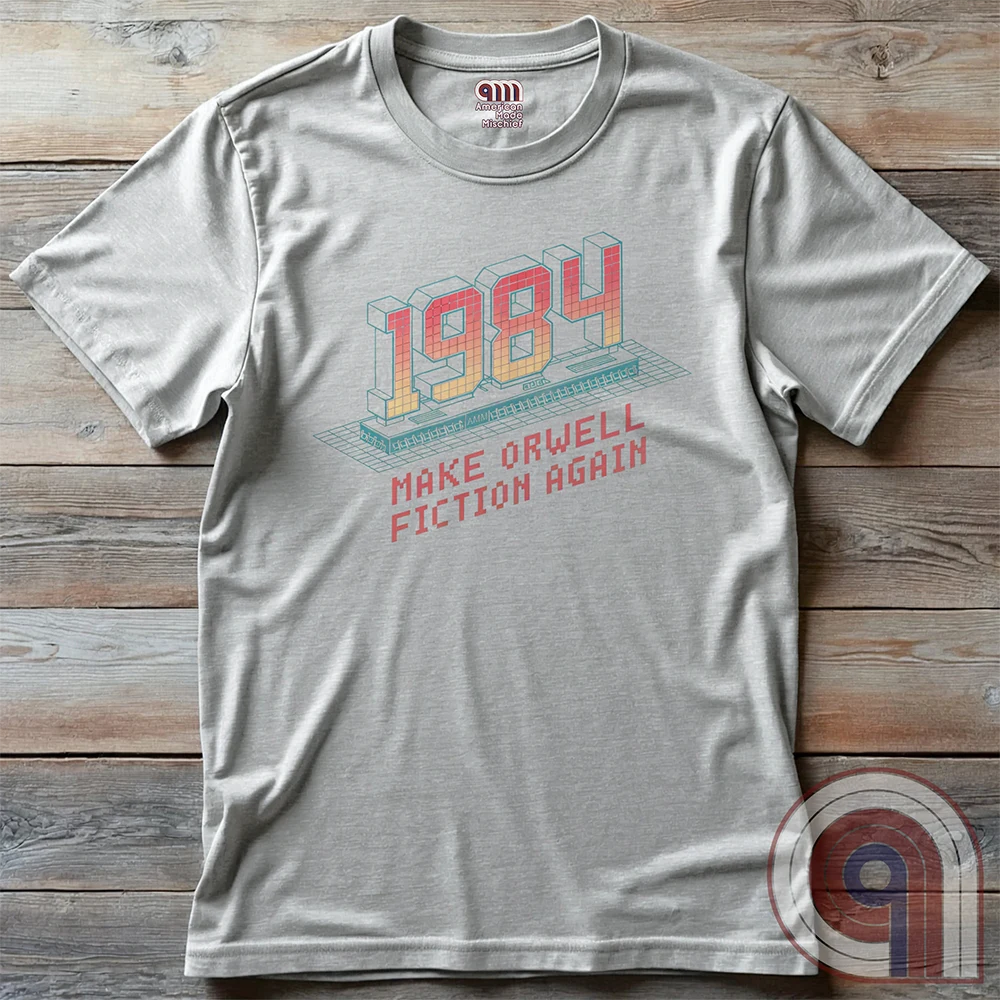 Light ash grey T-shirt laid flat on wooden boards. Its retro-computer graphic pairs pink-to-yellow 3D “1984” numbers with a turquoise keyboard and the pixelated line “MAKE ORWELL FICTION AGAIN”.