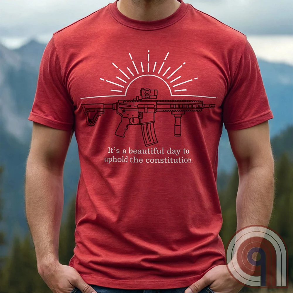 Red T-shirt on a model featuring a dark line-art AR-style rifle across the chest, white sunrise rays behind it, and the small printed sentence “It’s a beautiful day to uphold the constitution.”