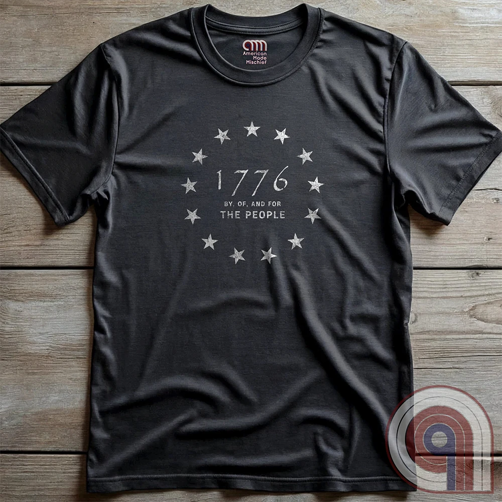Charcoal T-shirt laid flat on wood, showing a thirteen-star ring framing the year “1776” in vintage script, with “BY, OF, AND FOR THE PEOPLE” centered beneath it.