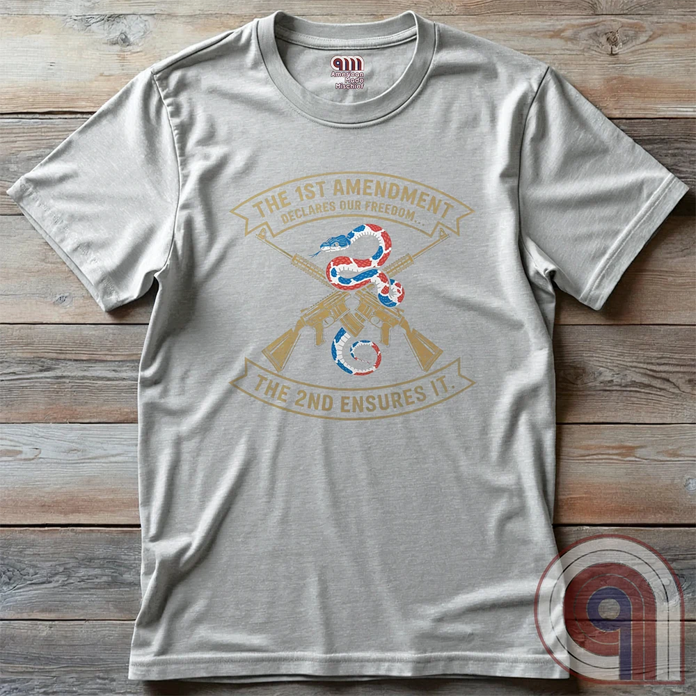 Light ash grey shirt laid flat on wood with two gold crossed rifles behind a flag-colored snake, surrounded by “THE 1ST AMENDMENT DECLARES OUR FREEDOM...” above and “THE 2ND ENSURES IT” below.