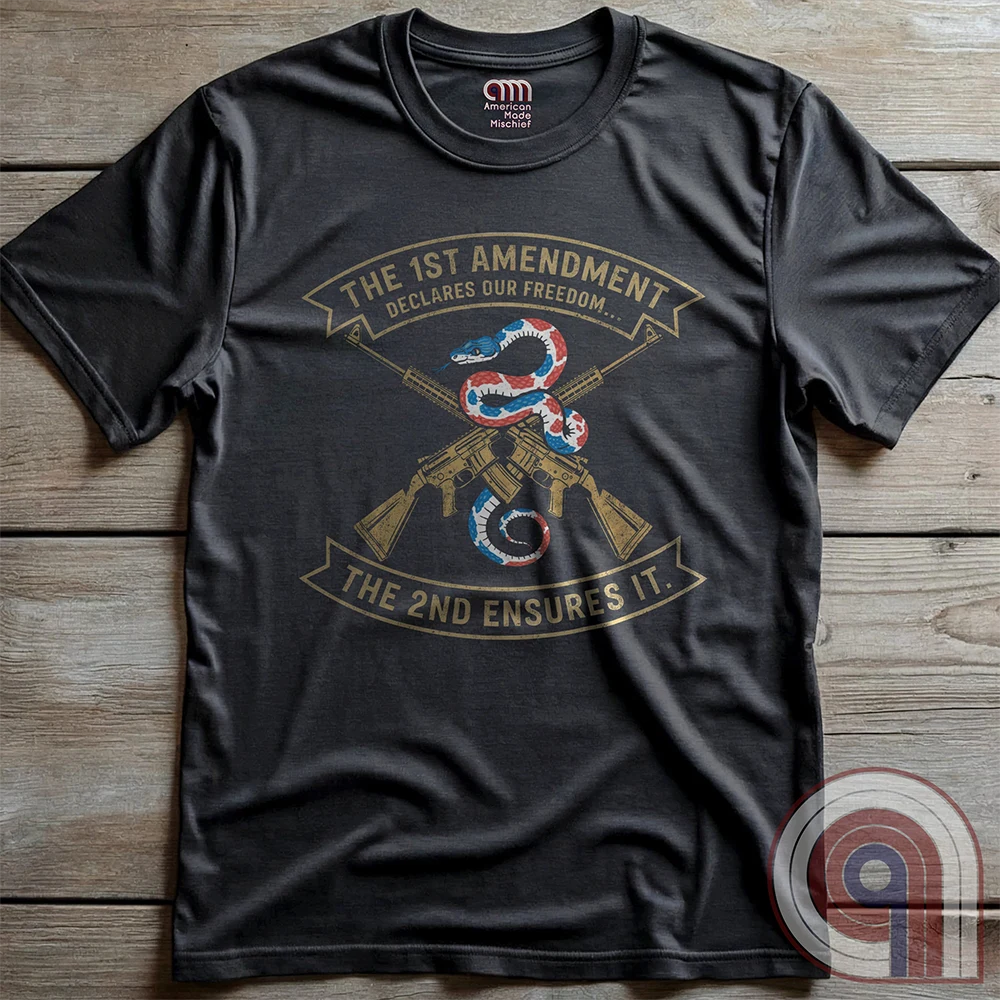Flat-lay charcoal tee on wood, with a red, white and blue rattlesnake curled over crossed gold rifles. The arched ribbons read “THE 1ST AMENDMENT DECLARES OUR FREEDOM...” and “THE 2ND ENSURES IT”.