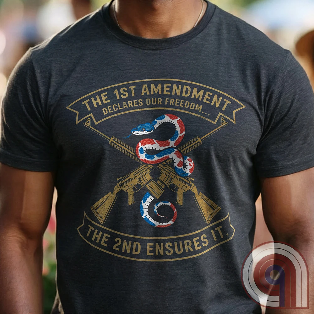 A charcoal T-shirt worn by a model shows a red, white and blue rattlesnake winding over crossed gold rifles, flanked by banners reading “THE 1ST AMENDMENT DECLARES OUR FREEDOM...” and “THE 2ND ENSURES IT”.