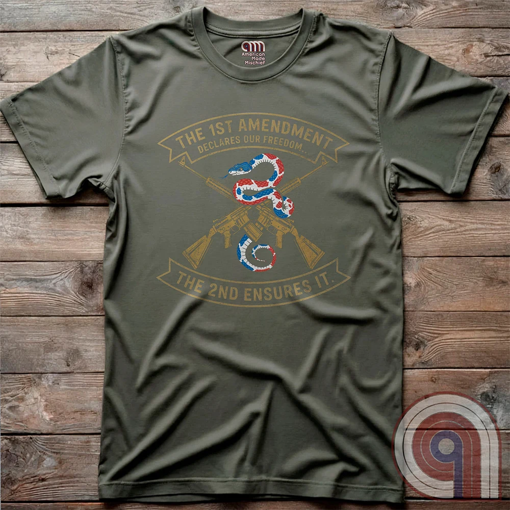 Olive green T-shirt photographed flat on wood, with gold rifles crossed behind a patriotic snake and two printed ribbons reading “THE 1ST AMENDMENT DECLARES OUR FREEDOM...” and “THE 2ND ENSURES IT”.