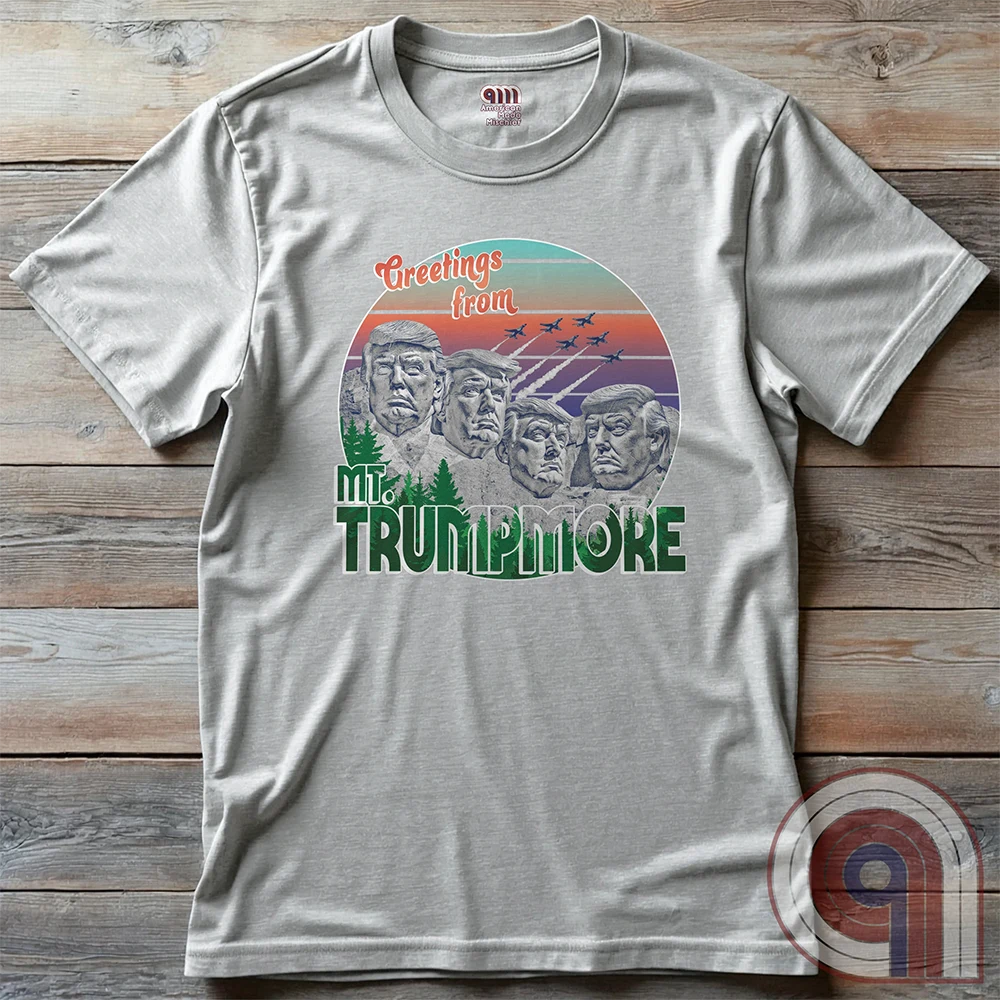 Light ash grey Mt. Trumpmore shirt arranged flat on wood. A retro sunset and small jets rise behind four carved Trump-like faces, with orange “Greetings from” script and green “TRUMPMORE” lettering.