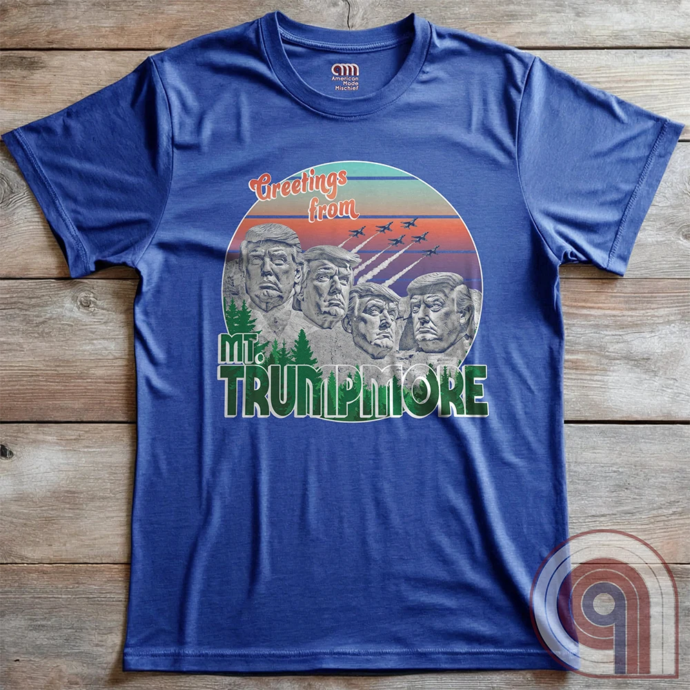 Heather blue T-shirt laid flat on wood, displaying the full vintage travel-souvenir spoof: “Greetings from Mt. TRUMPMORE,” four sculpted Trump likenesses and jet silhouettes.