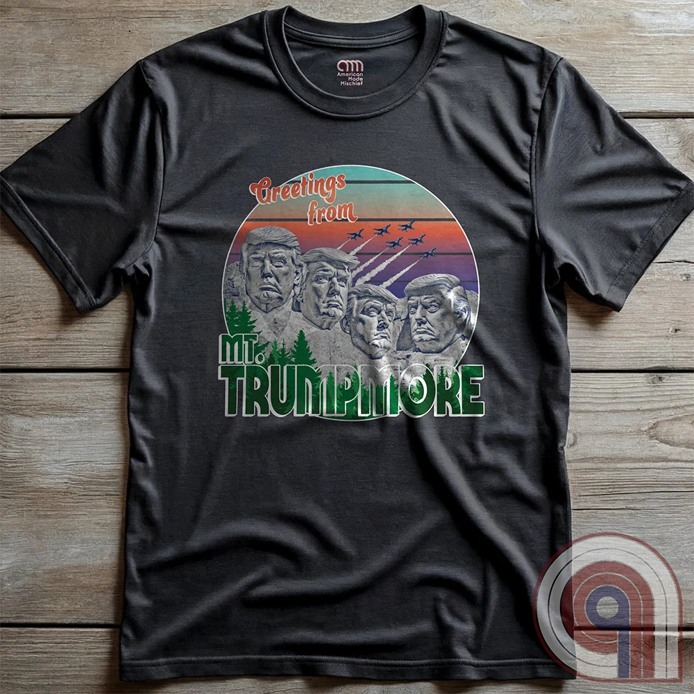 Charcoal grey shirt displayed flat on wood, featuring green “TRUMPMORE” lettering beneath a playful mountain monument of four Donald Trump faces and a vintage sunset with jets.