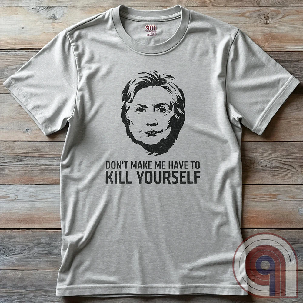 Light ash grey shirt laid flat on wooden boards, featuring a black stencil portrait of Hillary Clinton and the dark political-parody line “DON'T MAKE ME HAVE TO KILL YOURSELF”.