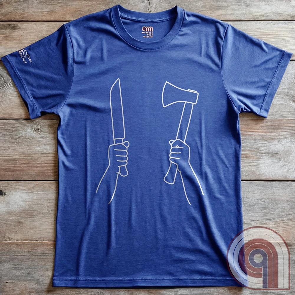 Blue T-shirt photographed flat on wood, showing the high-contrast paired fists holding a long knife and an upright hatchet.