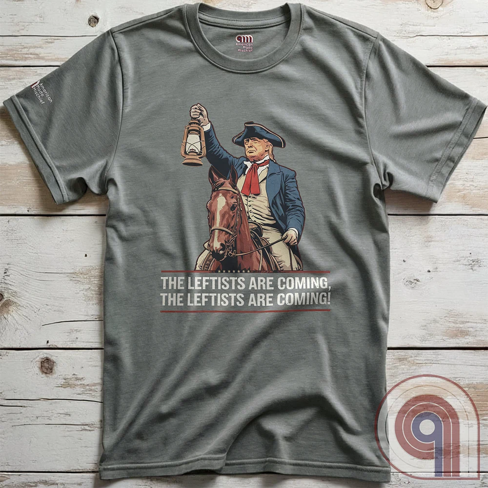 Heather grey Trump-as-Paul-Revere graphic T-shirt laid flat against wood, featuring a rider in a dark coat, red neckwear and broad hat on a brown horse above the repeated leftists warning.