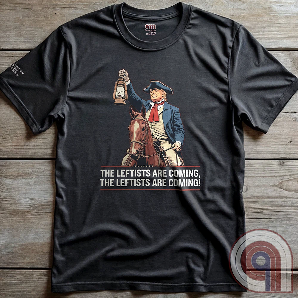 Charcoal grey T-shirt laid flat on wooden boards, with the historical spoof of a mounted Trump raising a lantern and the two-line “THE LEFTISTS ARE COMING, THE LEFTISTS ARE COMING!” slogan.