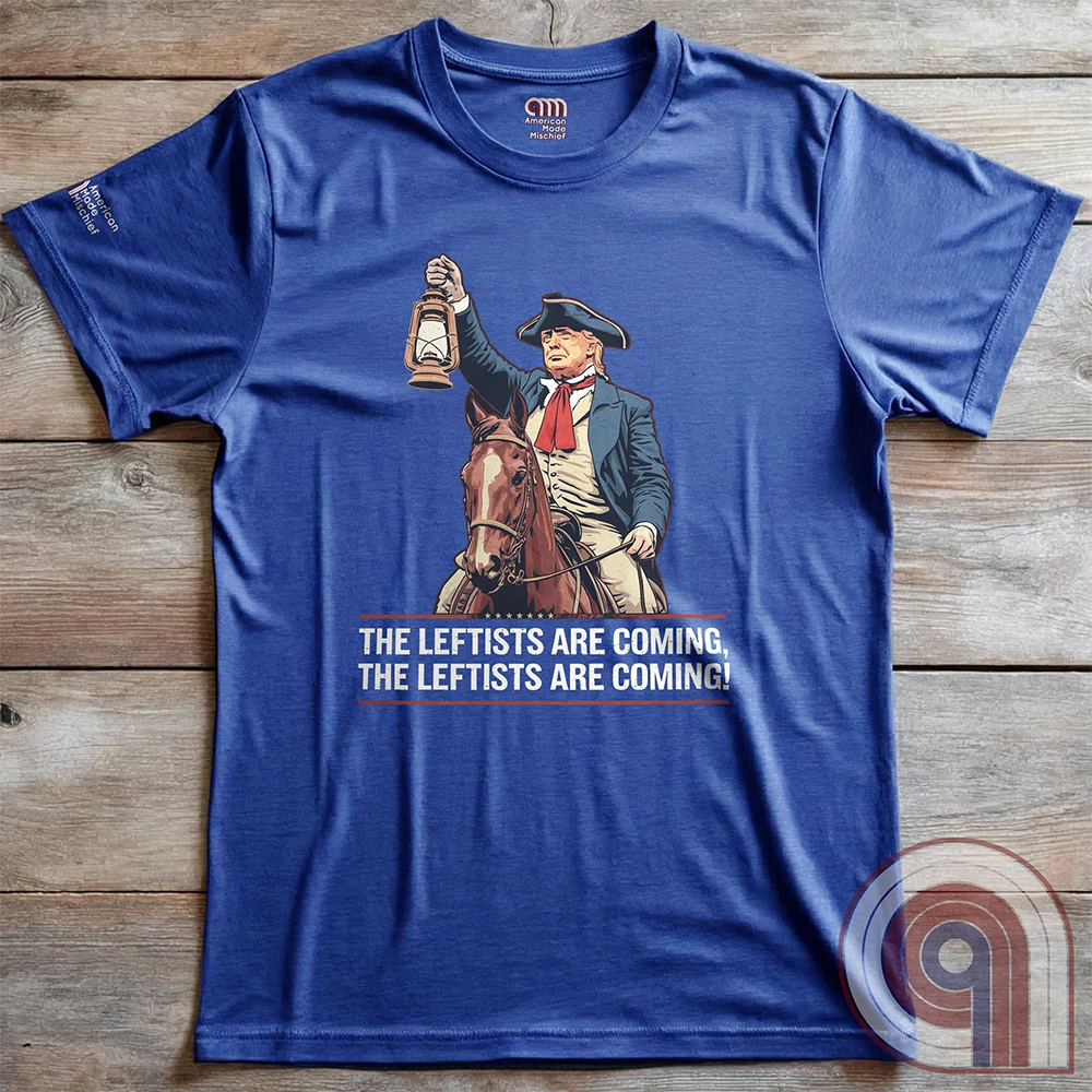 Heather blue T-shirt laid flat on wooden planks, with a vintage illustration of Trump as a lantern-raising horseback messenger and the repeated cry “THE LEFTISTS ARE COMING, THE LEFTISTS ARE COMING!”