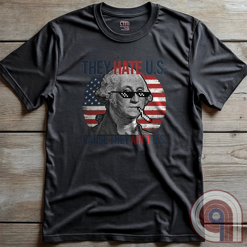 Charcoal T-shirt laid flat on wooden boards, displaying the complete George Washington and American flag parody in red, white and blue, with pixelated sunglasses, a cigarette and “THEY HATE U.S. ’CAUSE THEY AIN’T U.S.”