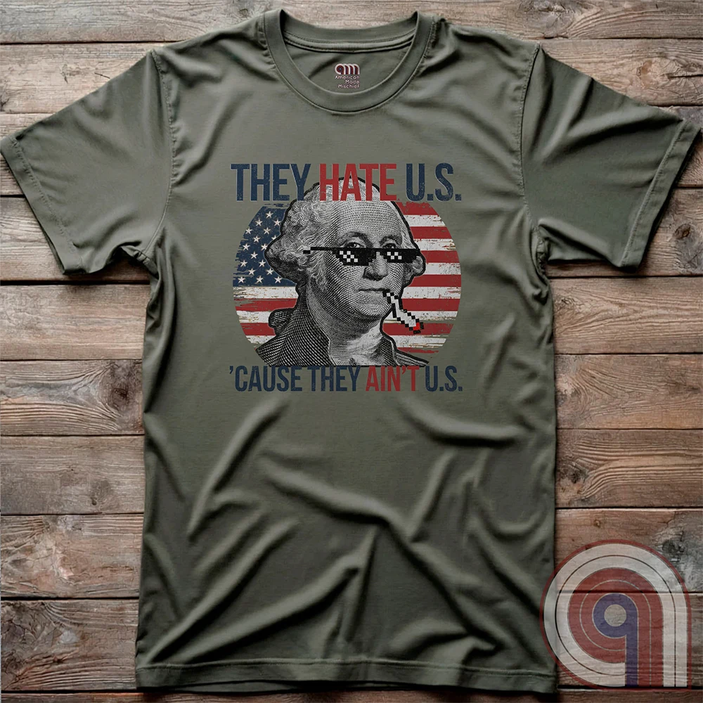 Flat-lay olive-green graphic tee on rustic wood, featuring George Washington in 8-bit-style shades with a cigarette, backed by red-and-white stripes and white stars. The bold patriotic meme reads “THEY HATE U.S. ’CAUSE THEY AIN’T U.S.”