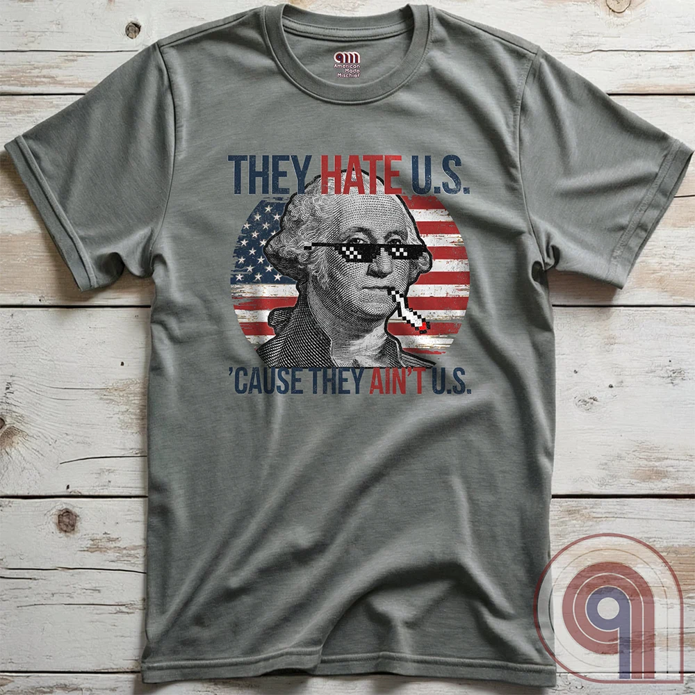 Heather-gray T-shirt displayed front-up on wooden planks, its George Washington thug-life parody centered over a faded U.S. flag. The print pairs pixelated sunglasses and a cigarette with the line “THEY HATE U.S. ’CAUSE THEY AIN’T U.S.”