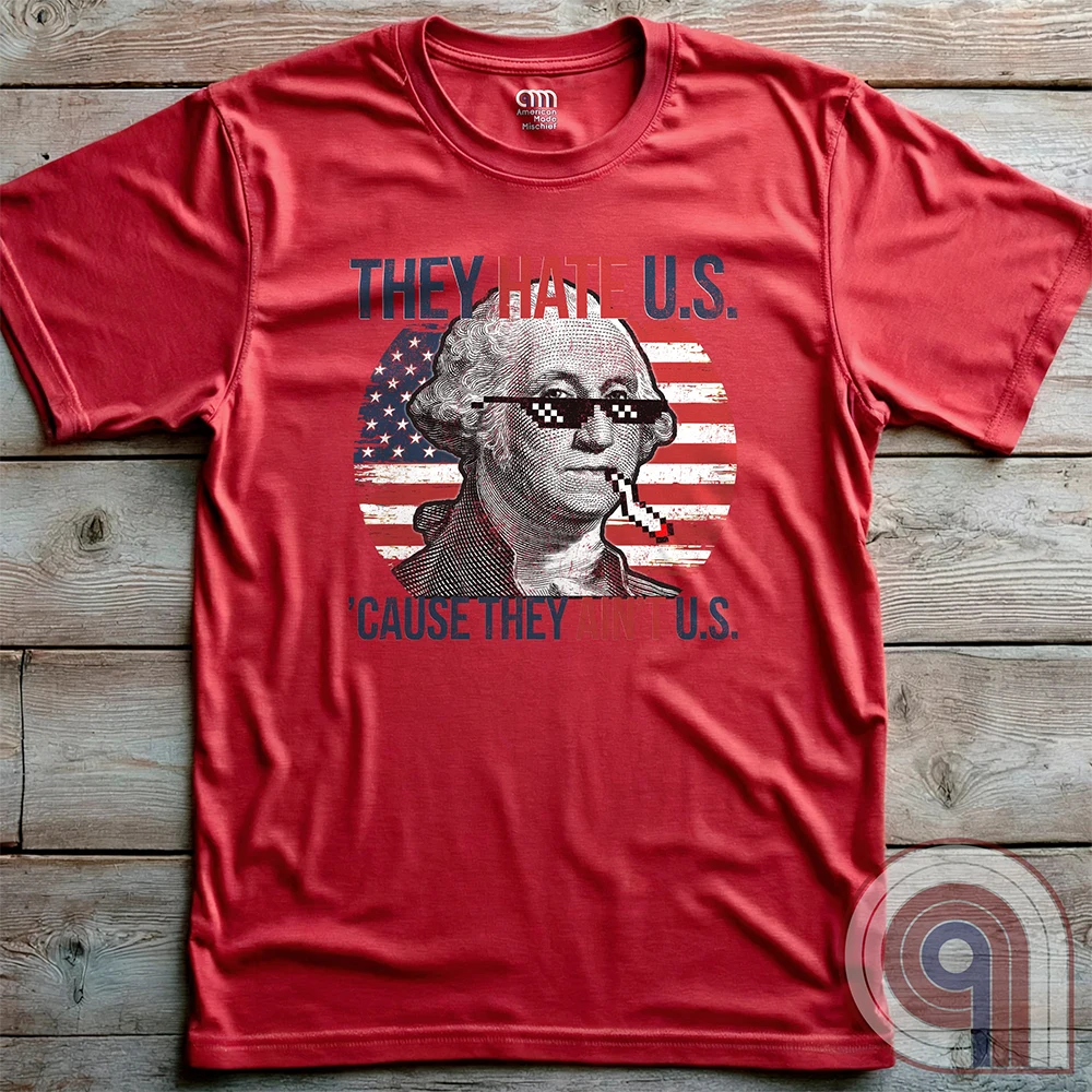 Red T-shirt photographed flat on a wooden surface; the chest print combines a monochrome George Washington portrait in pixel sunglasses with an American flag and the taunting wordplay “THEY HATE U.S. ’CAUSE THEY AIN’T U.S.”