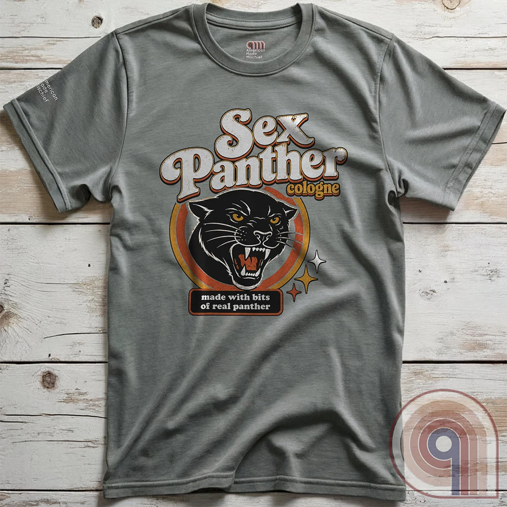 Heather-gray T-shirt photographed flat on wood, featuring the retro “Sex Panther cologne” logo, snarling black cat with bared teeth, orange concentric badge and starbursts. The fictional authenticity tag reads “made with bits of real panther.”