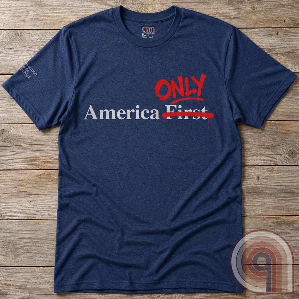 A heather navy T-shirt arranged flat on wooden planks, featuring pale “America First” text with “First” crossed out and “ONLY” written in red.