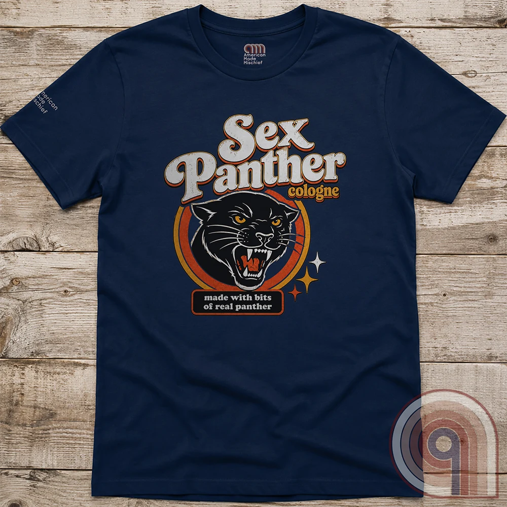 Navy T-shirt displayed flat on wood, with retro cream “Sex Panther cologne” typography over a snarling black panther in an orange emblem. A small seal jokes “made with bits of real panther,” echoing the Anchorman cologne gag.