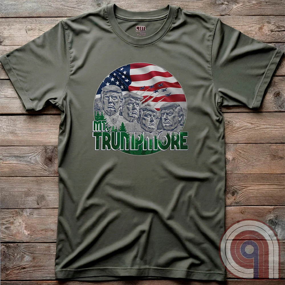 Olive green shirt laid flat on wood, featuring the “Mt. TRUMPMORE” design: a patriotic Mount Rushmore spoof with four stone-faced Trump portraits beneath Old Glory.