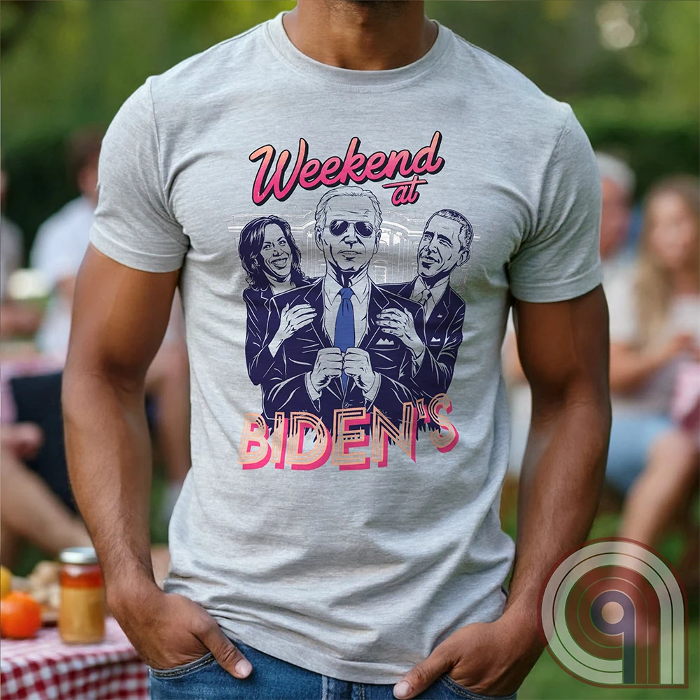 Light heather gray T-shirt worn by a model, featuring “Weekend at BIDEN’S,” a Weekend at Bernie’s movie parody portraying Joe Biden in sunglasses propped up by caricatures of Kamala Harris and Barack Obama, in retro purple and pink.