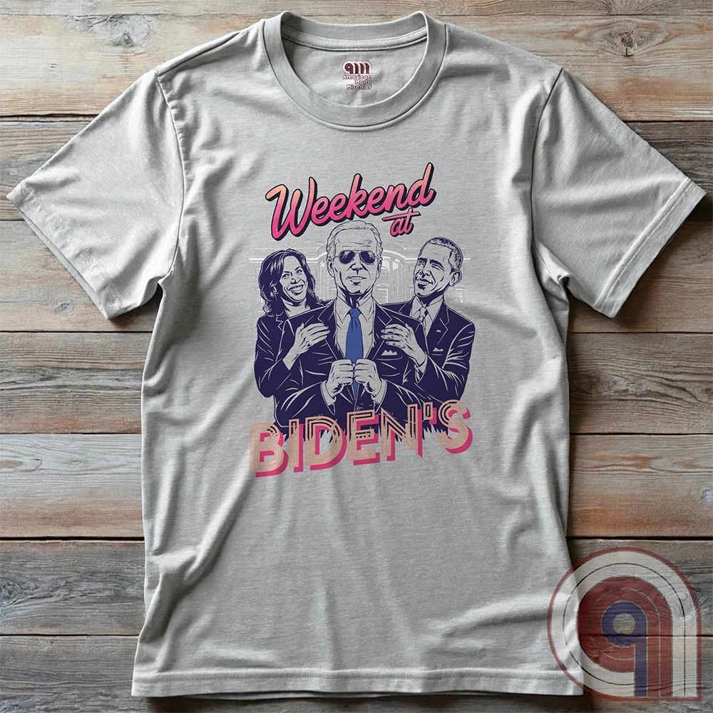 Ash-gray T-shirt laid flat on wood, displaying the full “Weekend at BIDEN’S” comedy parody: Joe Biden in sunglasses between caricatures of Kamala Harris and Barack Obama, who appear to hold him upright.