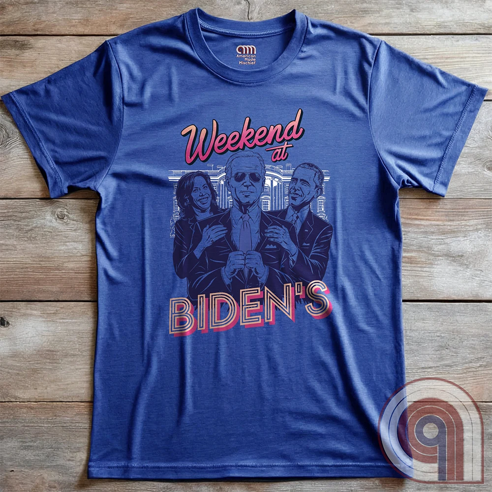 Blue T-shirt laid flat on wood, with the “Weekend at BIDEN’S” parody of Weekend at Bernie’s: purple caricatures of Joe Biden, Kamala Harris and Barack Obama framed by pink script and block lettering.