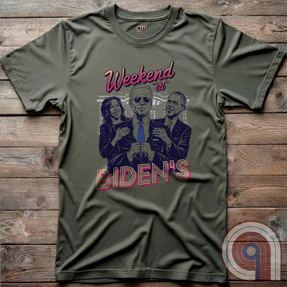 Olive green T-shirt laid flat on planks, printed with pink “Weekend at BIDEN’S” typography and a purple-toned political comedy parody portraying Joe Biden held between Kamala Harris and Barack Obama.