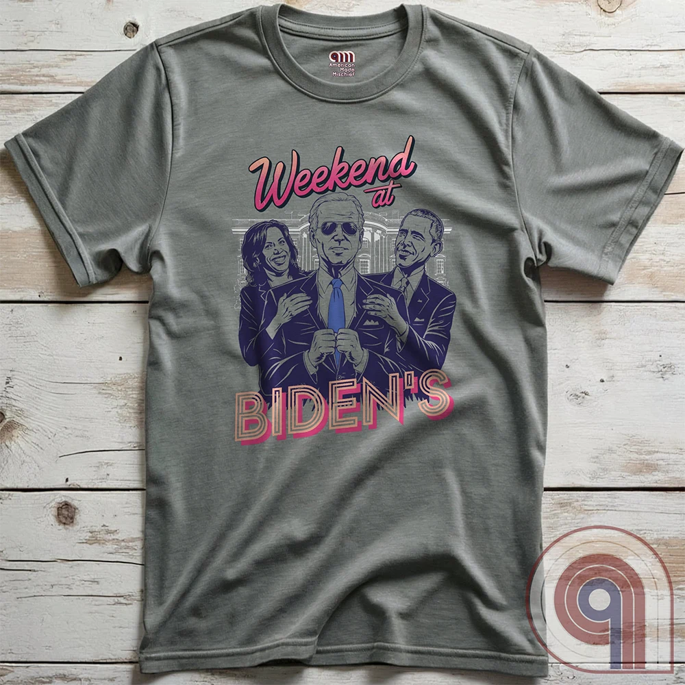 Heather gray T-shirt flat on weathered wood, featuring a Weekend at Bernie’s-style “Weekend at BIDEN’S” satire with Joe Biden in sunglasses between Kamala Harris and Barack Obama, their hands on his shoulders.